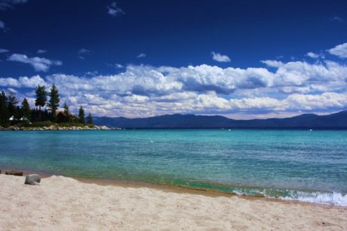 A serene beach scene with turquoise water, sandy shore, and a backdrop of clouds and mountains under a blue sky.