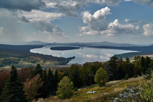 A serene landscape featuring a calm lake surrounded by lush forests and distant mountains under a cloudy sky.
