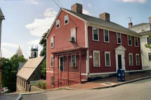 A red historic house on a sloped street, with a church steeple and a postal box nearby under a blue sky.