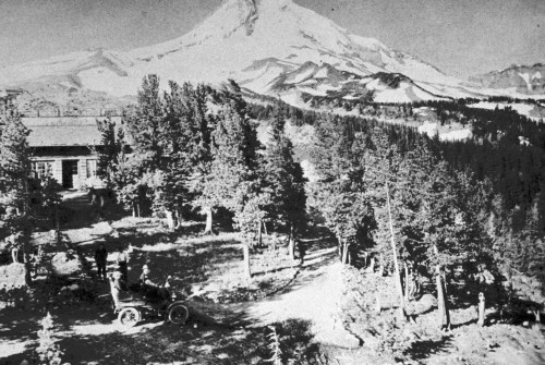 Black and white image of a vintage car near a forested area with a mountain in the background.