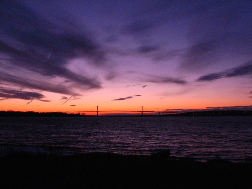 A serene sunset over a body of water, with a bridge silhouetted against vibrant purple and orange skies.