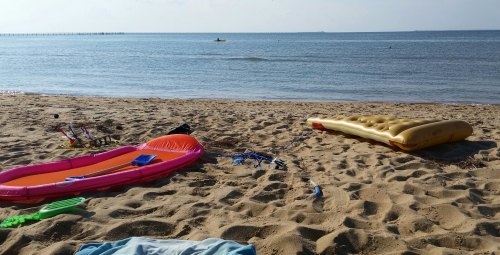 Colorful inflatable rafts on a sandy beach with calm water and a distant figure kayaking.