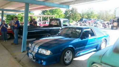 A blue sports car with racing stripes parked under a shelter, surrounded by people and classic vehicles.