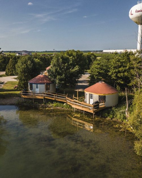 Aerial view of two round cabins on stilts by a pond, surrounded by trees and a water tower in the background.