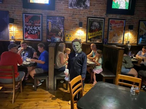 A person in a Michael Myers costume stands in a restaurant filled with diners, surrounded by vintage horror movie posters.