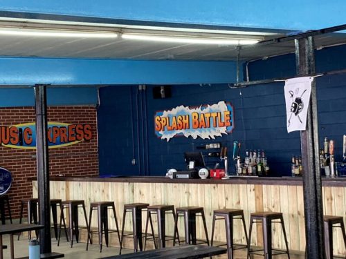 Interior of a bar featuring a "Splash Battle" sign, wooden bar, and high stools against a blue wall.