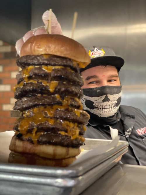 A chef in a skull mask holds a towering burger stacked with multiple patties and cheese, set in a kitchen.