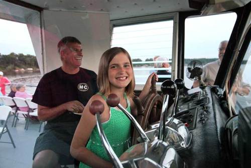 A girl smiles while steering a boat, with an adult behind her. The setting is relaxed and cheerful.