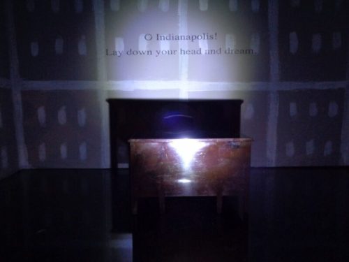 A dimly lit room features a small table with a spotlight and the text: "O Indianapolis! Lay down your head and dream."