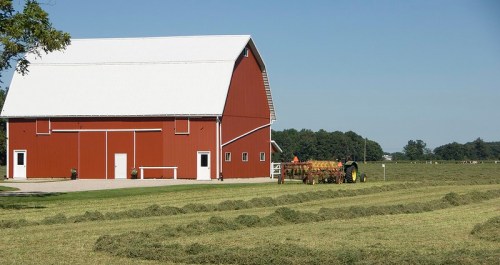 A red barn stands beside freshly cut hay in a sunny field, with a tractor working nearby under a clear blue sky.