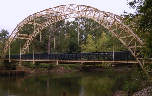 A yellow arch bridge spans a calm river, surrounded by lush green trees and foliage.
