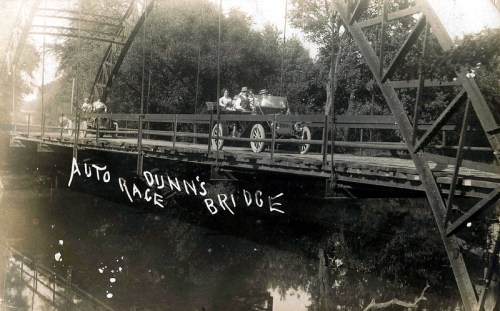 Historic photo of a vintage car on Dunn's Bridge, with people nearby and "Auto Race" written on the bridge.