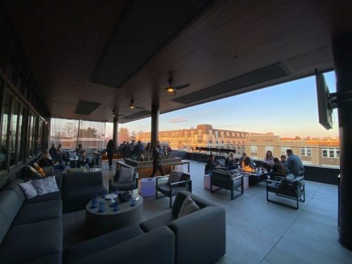 A rooftop lounge with seating and fire pits, filled with people enjoying the sunset over city buildings.