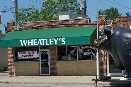 A storefront with a green awning reading "Wheatley's" and a large cow statue outside, advertising a fish fry.