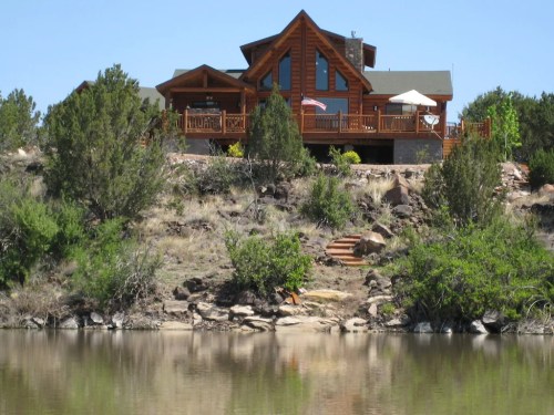 A large log cabin on a hillside overlooking a calm lake, surrounded by greenery and blue skies.