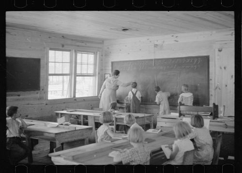 A teacher writes on a chalkboard while students observe in a vintage classroom with wooden desks and a rustic interior.