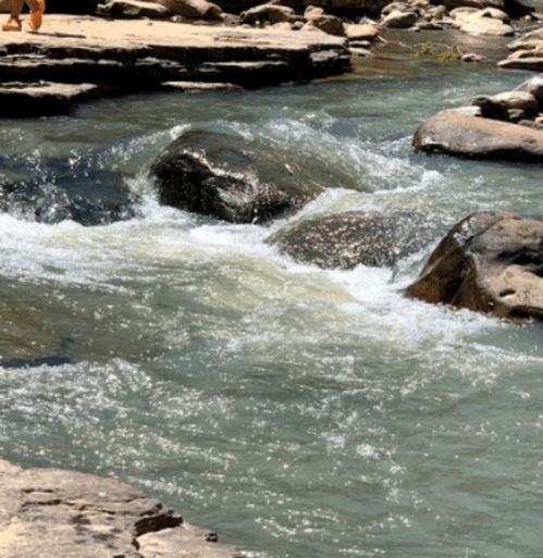 A flowing river with clear water and smooth rocks, surrounded by a natural landscape.