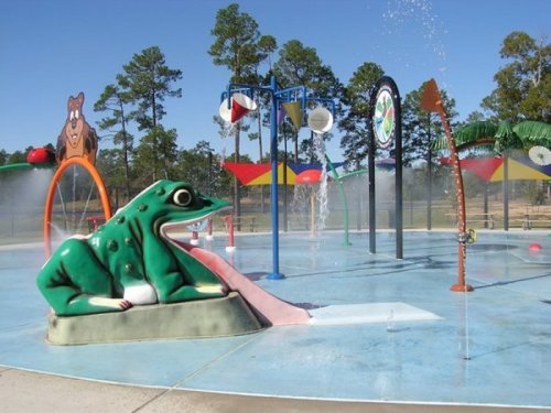 A colorful splash pad featuring a large frog sculpture and various water features, surrounded by trees and blue sky.