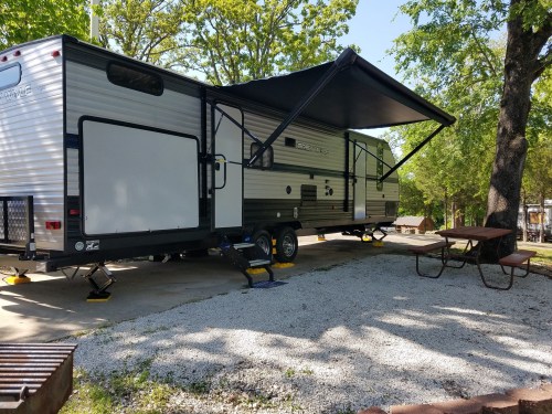 A parked RV with an extended awning, surrounded by trees and a gravel area with a picnic table.