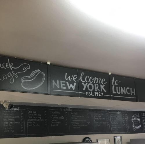 Chalkboard sign reading "Welcome to New York est. 1927" above a menu board in a casual dining setting.