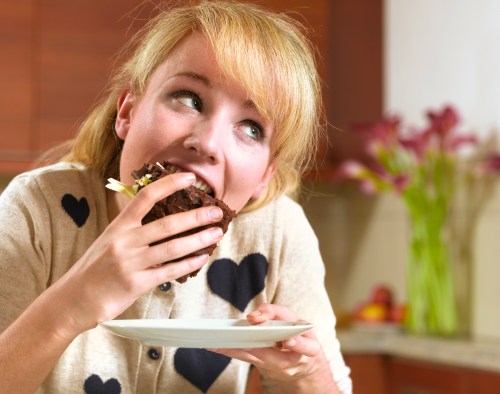 A woman with blonde hair is about to take a big bite of a chocolate dessert while sitting in a kitchen.