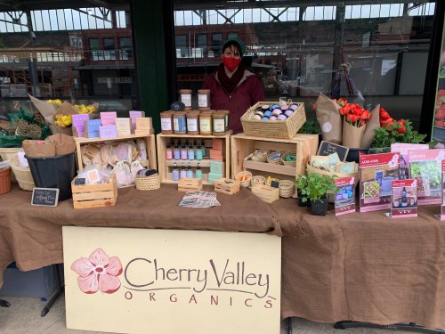 A vendor at a market stand displays organic products, including flowers, jars, and plants, with a sign for Cherry Valley Organics.