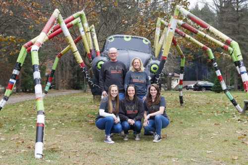 A family poses in front of a large spider sculpture made of colorful materials in a park setting.