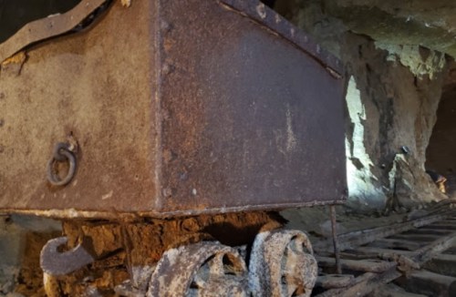 Rusty mining cart on tracks inside a dimly lit cave, with rocky walls in the background.