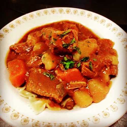 A bowl of hearty beef stew with vegetables, garnished with herbs, served over noodles in a decorative dish.