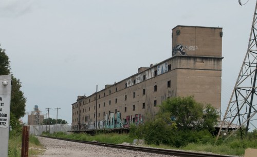 Abandoned industrial building with graffiti, located near railway tracks and surrounded by overgrown vegetation.