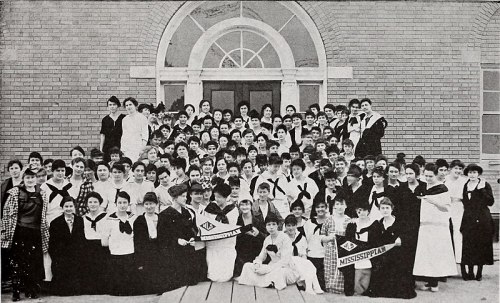 A large group of women in early 1900s attire poses in front of a brick building, holding banners for Mississippi.