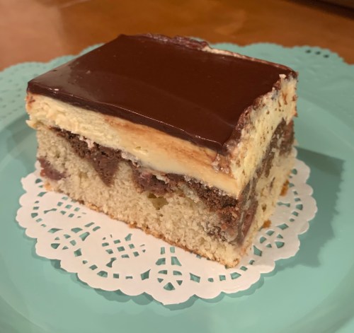 A slice of layered cake with chocolate frosting on top, sitting on a decorative doily on a teal plate.