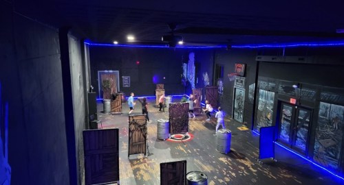 A dimly lit indoor arena with blue lights, featuring players engaged in a laser tag game among obstacles.
