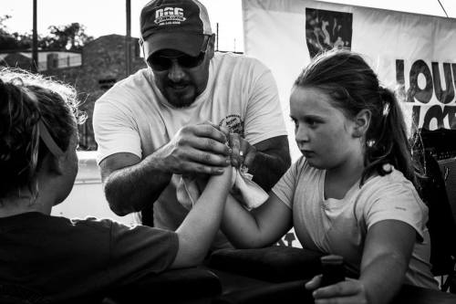 A man assists two girls in an arm wrestling match at an outdoor event, with a focused atmosphere.