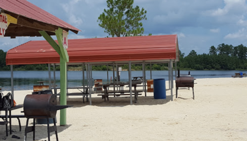 Beachside picnic area with red-roofed shelters, grills, and a calm lake in the background under a partly cloudy sky.