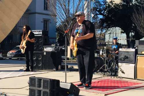 A band performs outdoors with a guitarist singing, a bassist, and amplifiers in the background under a clear blue sky.