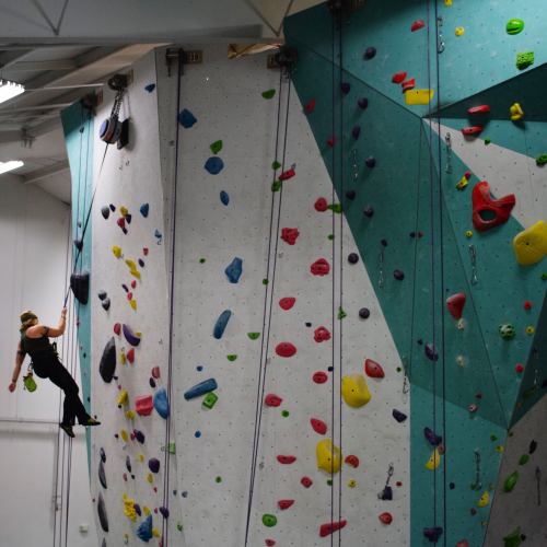 A climber ascends a colorful indoor rock wall, surrounded by various climbing holds and ropes.