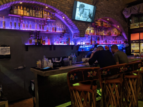 A dimly lit bar with colorful shelves of liquor, two patrons seated at the counter, and a vintage film playing on a screen.