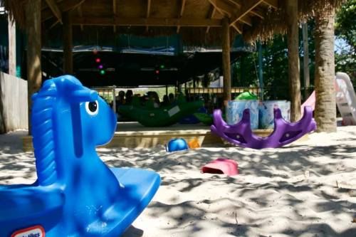 Colorful playground equipment, including a blue rocking horse, sits on sandy ground under a shaded structure.