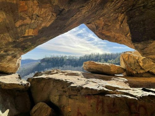 View from a cave opening, showcasing a scenic landscape with trees and a blue sky in the background.