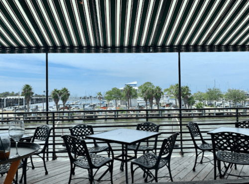 View from a restaurant patio overlooking a marina with boats and palm trees under a striped awning.