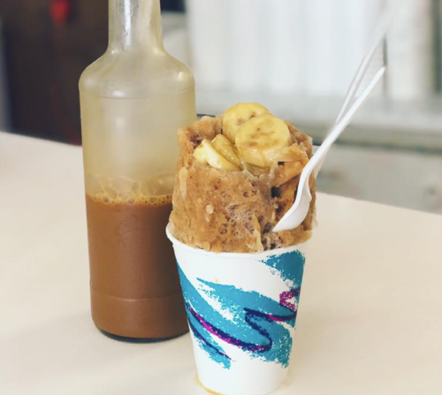 A cup of ice cream topped with banana slices, next to a bottle of syrup on a white surface.