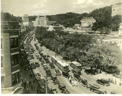 A busy street scene from the past, filled with vintage cars and people, surrounded by trees and buildings.