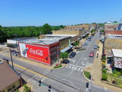 Aerial view of a small town street featuring a Coca-Cola mural, shops, and parked cars under a clear blue sky.