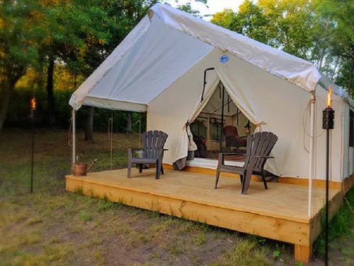 A cozy tent on a wooden deck, surrounded by trees, with two chairs on the porch and torches nearby.