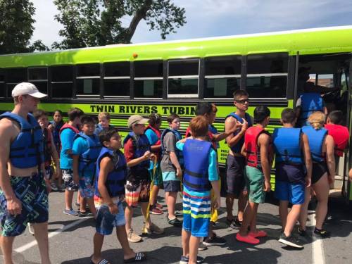 A group of children in life jackets boarding a bright green bus labeled "Twin Rivers Tubing" on a sunny day.