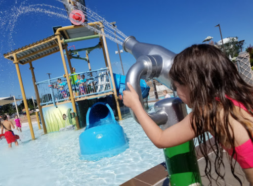 A child plays with a water spray feature at a splash pad, with a colorful play structure and other kids in the background.
