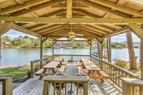 A lakeside pavilion with wooden picnic tables and a ceiling fan, overlooking a calm water view.