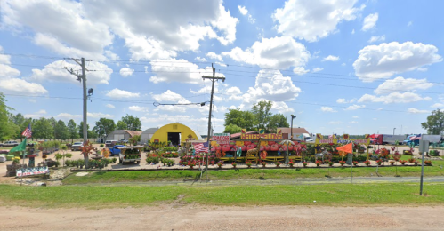 A colorful garden center with various plants and decorations, set against a bright blue sky with fluffy clouds.