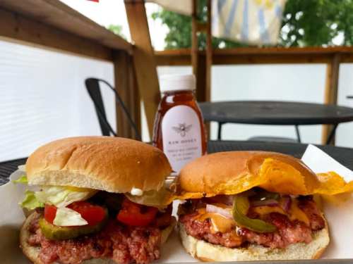 Two burgers with lettuce, tomato, pickles, and cheese, alongside a bottle of raw honey on a table.